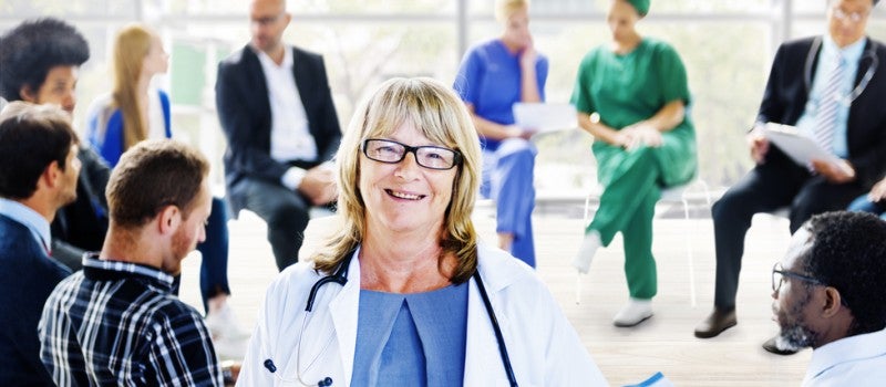 A female doctor smiling at the camera - behind her are other doctors in a meeting. 