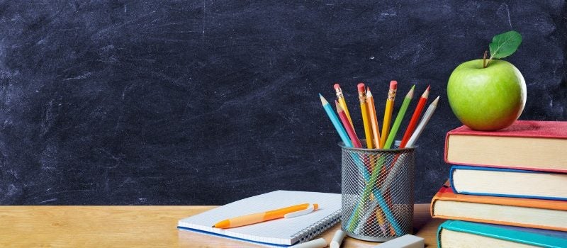 School items on a wooden desk: notepad, pencils, books and an apple. 
