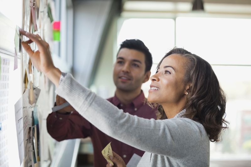 An SCU Online Learning Designer and academic stand in front of a white board, smiling and adding post-it notes to the board.