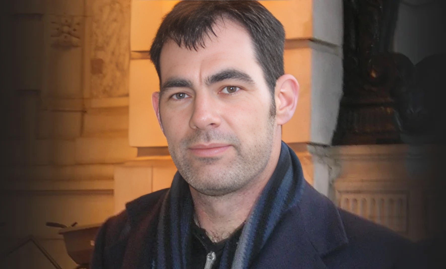 A man wearing a black jacket and scarf gazes directly at the camera, exuding confidence and style.