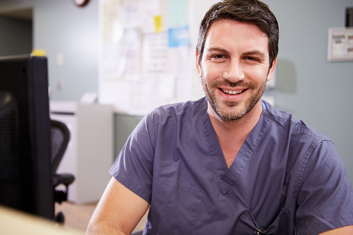A cheerful male mental health student wearing scrubs is seated at a desk, radiating warmth and confidence in his medical environment