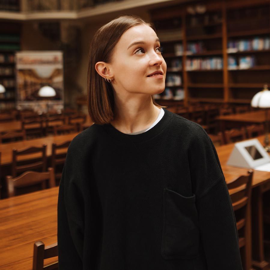A woman stands in a library, gazing upwards, surrounded by shelves filled with books.