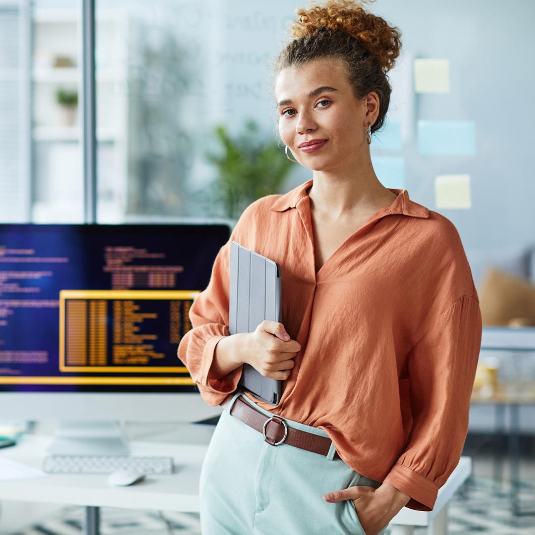 A professional woman in an office environment, holding a laptop and appearing focused on her tasks.