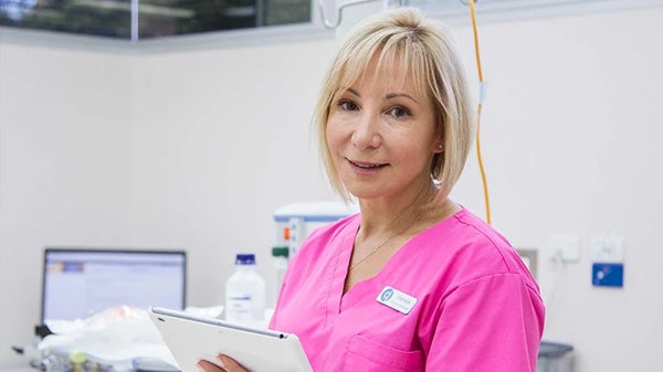 A woman in a pink scrub suit is holding a tablet, appearing focused and engaged in her work environment.
