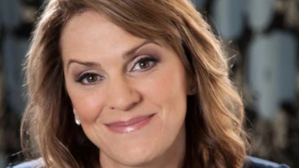 A woman with brown hair wearing a blue shirt, smiling and standing against a neutral background.