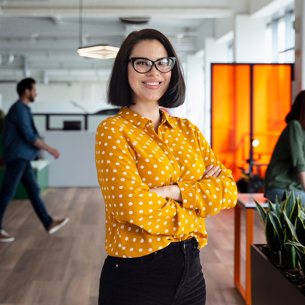 A woman wearing glasses stands in an office surrounded by colleagues engaged in conversation and collaboration.
