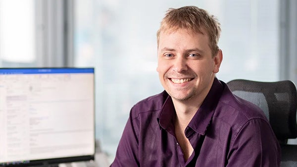 A man wearing a purple shirt is seated at a desk, focused on his work in a well-lit office environment.