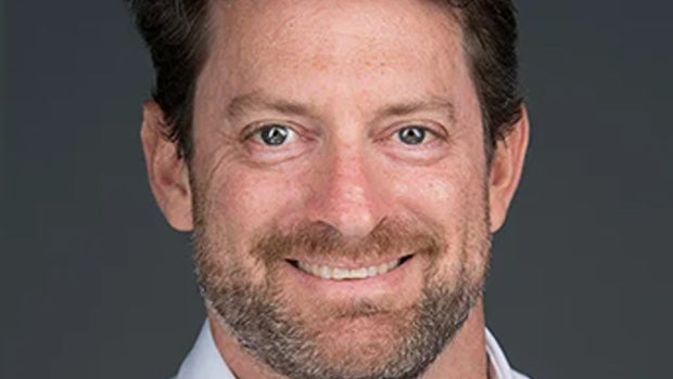 A bearded man wearing a white shirt, standing confidently with a neutral expression in a well-lit environment.