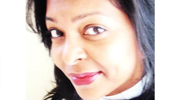 A woman with dark hair wearing a white shirt, standing confidently with a neutral expression against a plain background.