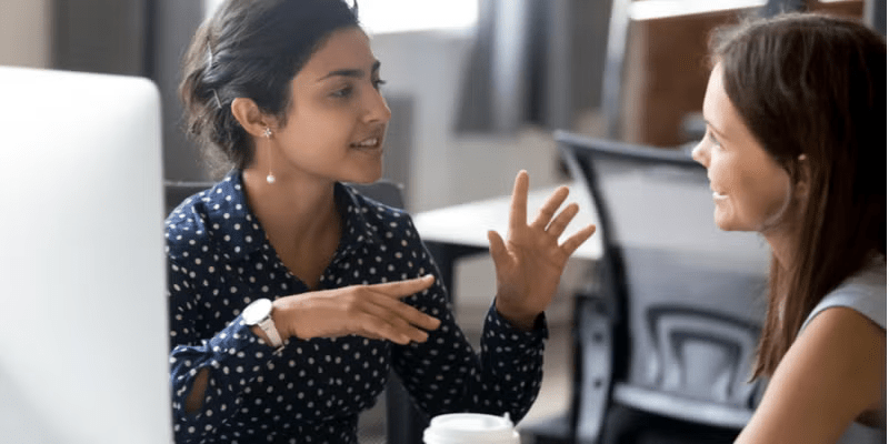 AN SCU Online MBA graduate is sitting at a desk, having an animated discussion with a co-worker.