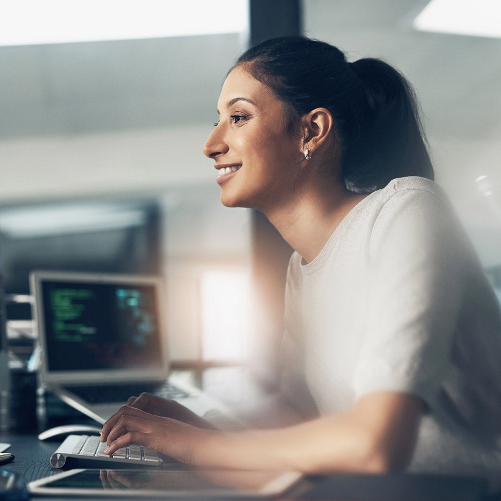 A woman seated at a desk, focused on her laptop, surrounded by a tidy workspace and soft lighting.
