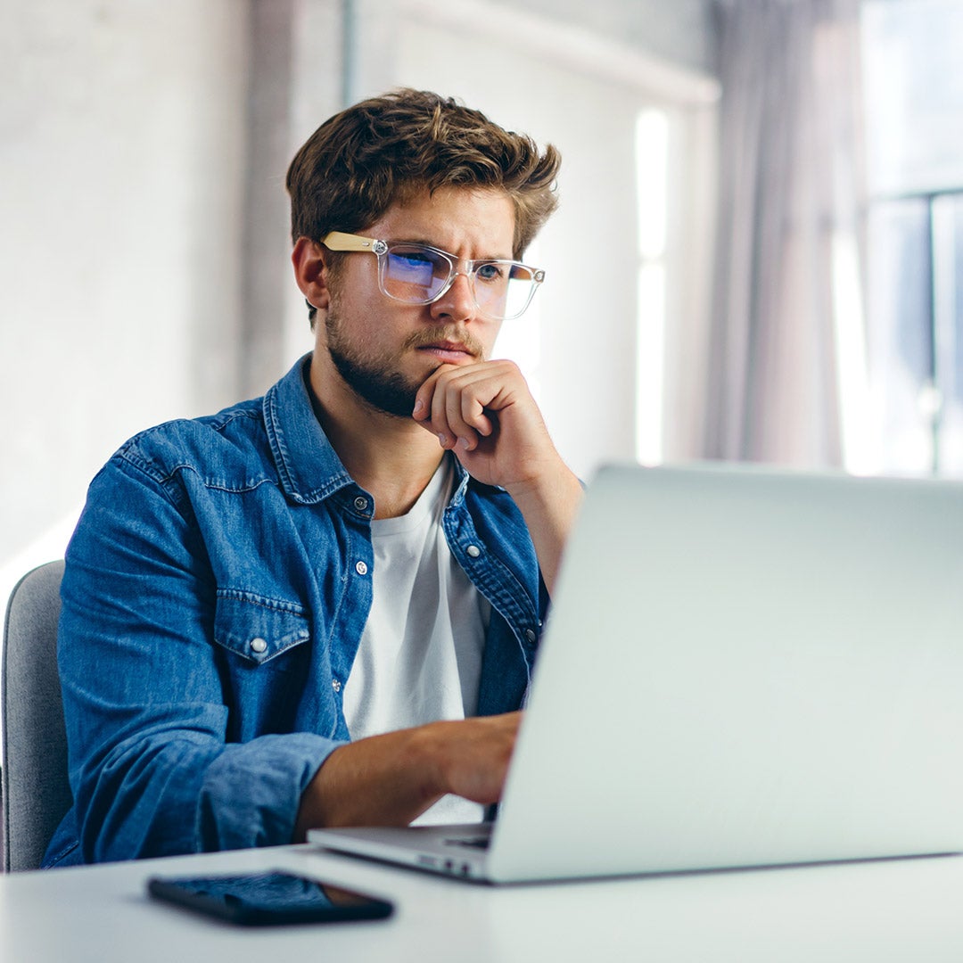 A man with glasses sits at a desk, focused on his laptop, surrounded by a tidy workspace.
