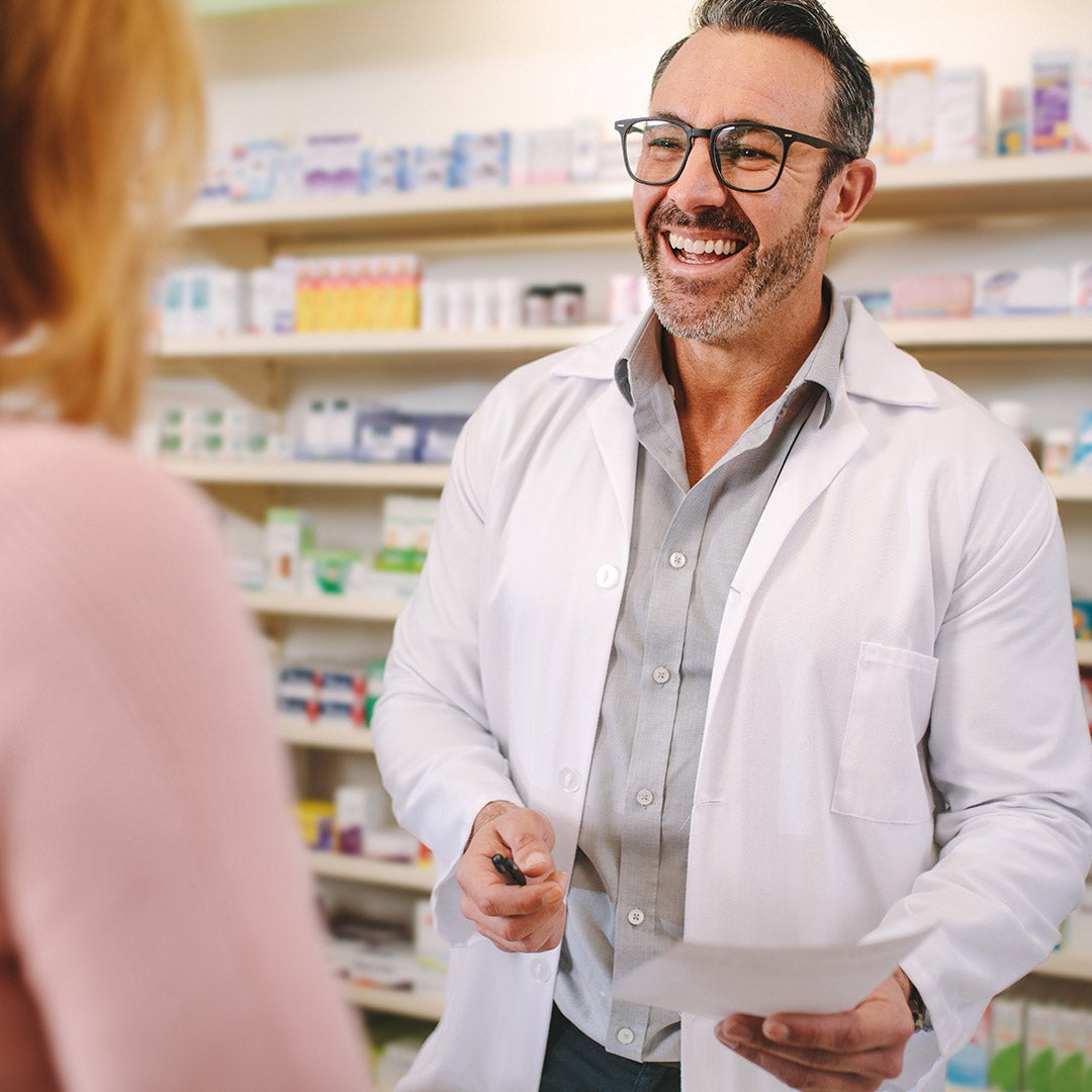 A man in a white coat stands before shelves filled with various medicine bottles and packages.