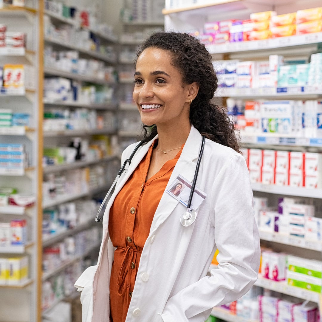 A smiling woman stands in a pharmacy shop, surrounded by shelves filled with various health products and medications.