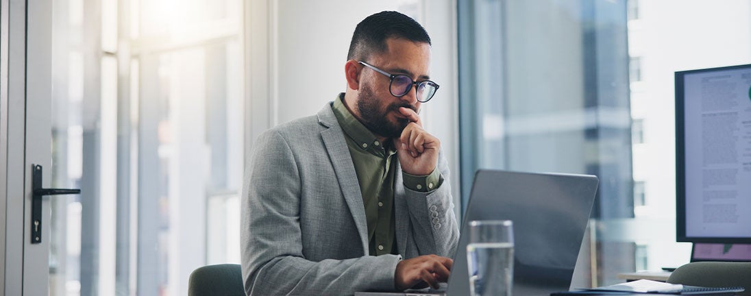 A man wearing glasses is seated at a desk, focused on his laptop while working intently.
