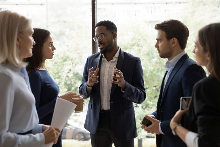 Five professionally dressed people stand in front of an office window having a discussion.