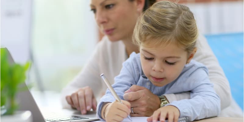 A working mother studying on her laptop. Her daughter is sat next to her drawing.