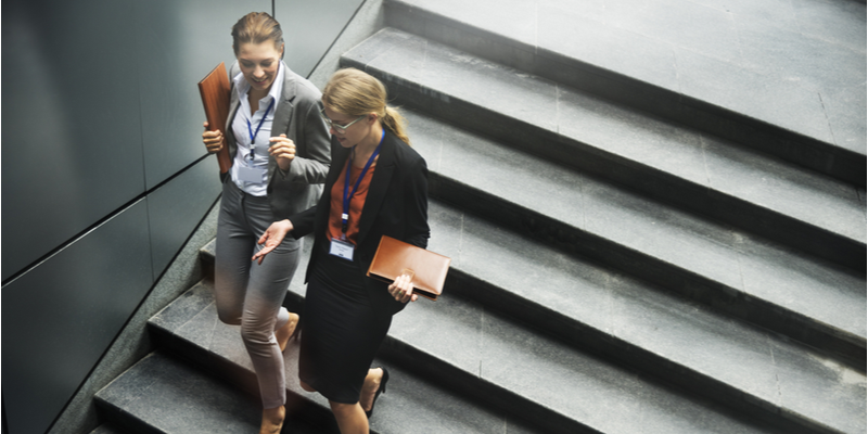 Two corporate business women chatting while walking down some stairs.