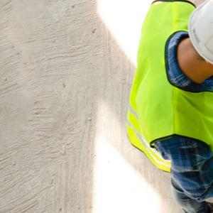 Three individuals in hard hats and safety vests examine blueprints at a construction site, focused on their task.