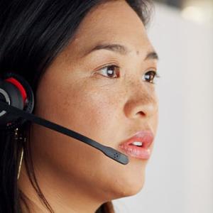 A woman in an office wearing a headset, engaged in a conversation, with a focused expression on her face.