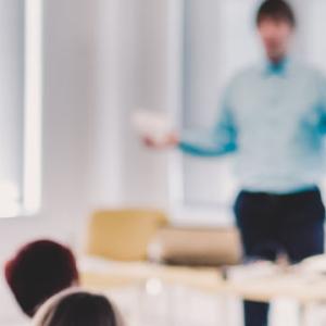 A man stands at the front, presenting to an engaged audience of diverse individuals seated in a conference room.