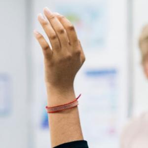 A woman stands in a classroom, raising her hands enthusiastically as she engages with her students.