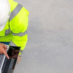 Three men in hard hats and safety vests collaborate over a laptop, focused on their work in a construction environment.