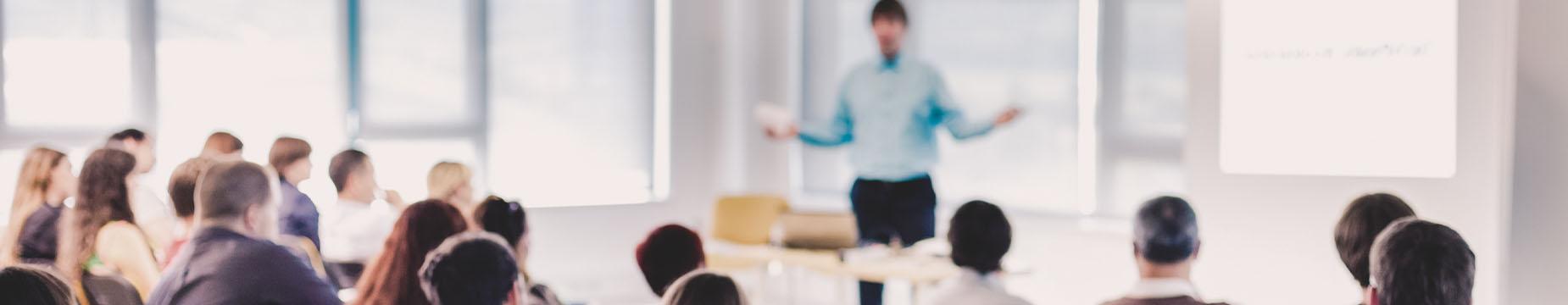 A man stands at the front, presenting to an engaged audience of diverse individuals seated in a conference room.