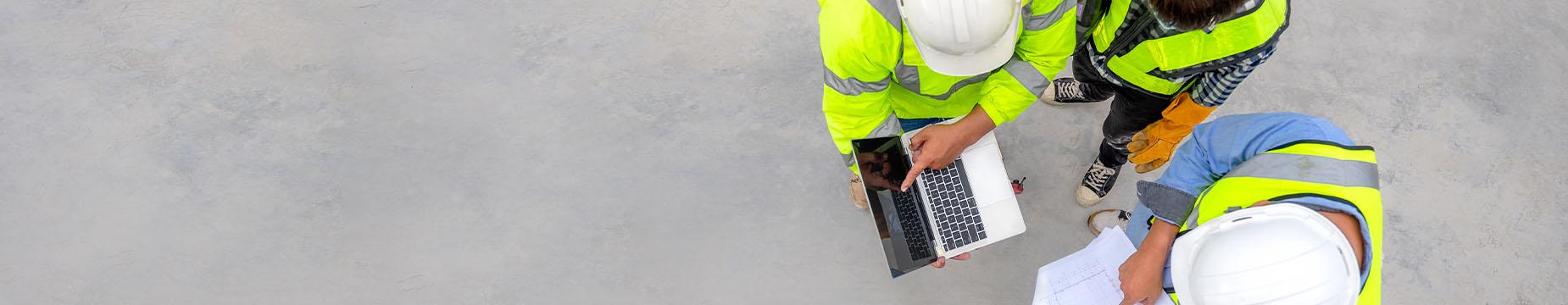 Three construction workers wearing yellow vests and hard hats collaborate on a building site, focused on their tasks.