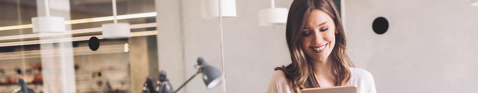 A woman stands in front of a desk, holding a tablet, engaged in a focused task or discussion.