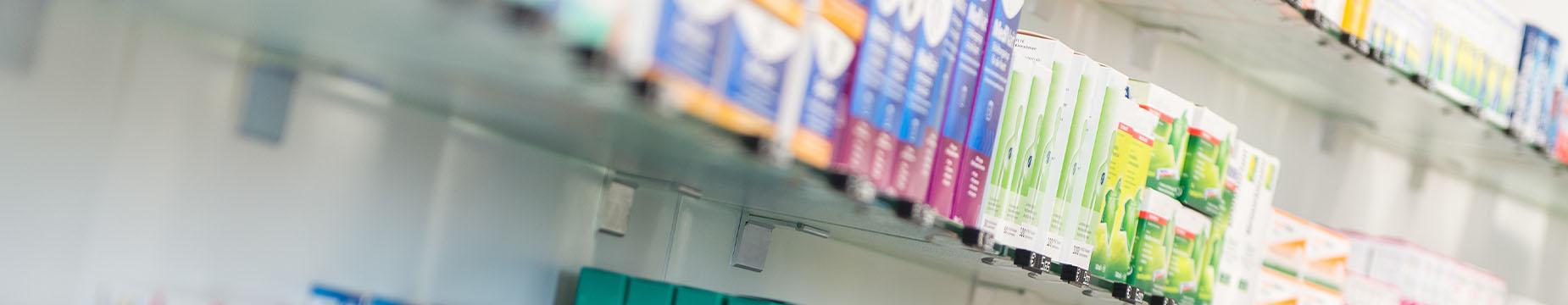 A man in a white coat stands before shelves filled with various medicine bottles and packages.