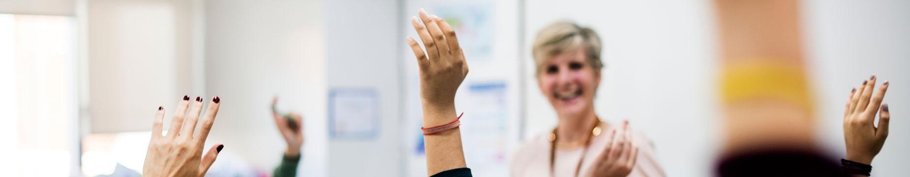 A woman stands in a classroom, raising her hands enthusiastically as she engages with her students.