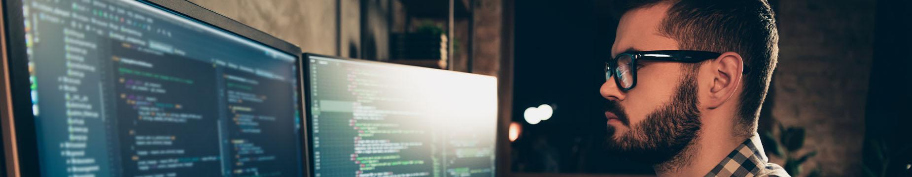 A man wearing glasses is focused on his work, utilising two computer screens for multitasking.