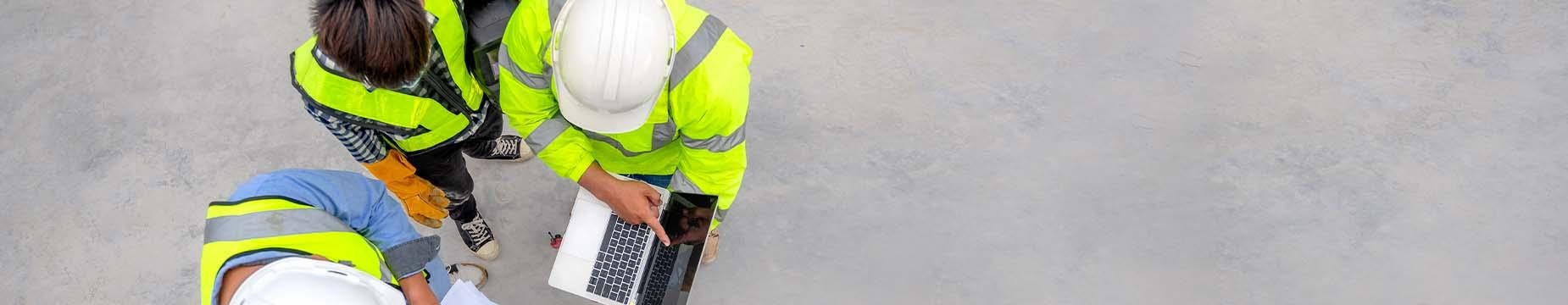 Three men in hard hats and safety vests collaborate over a laptop, focused on their work in a construction environment.