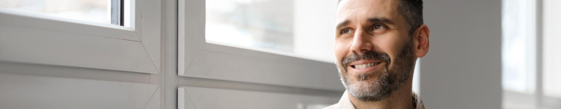 A bearded man wearing a watch stands thoughtfully in front of a large window, enjoying the view outside.