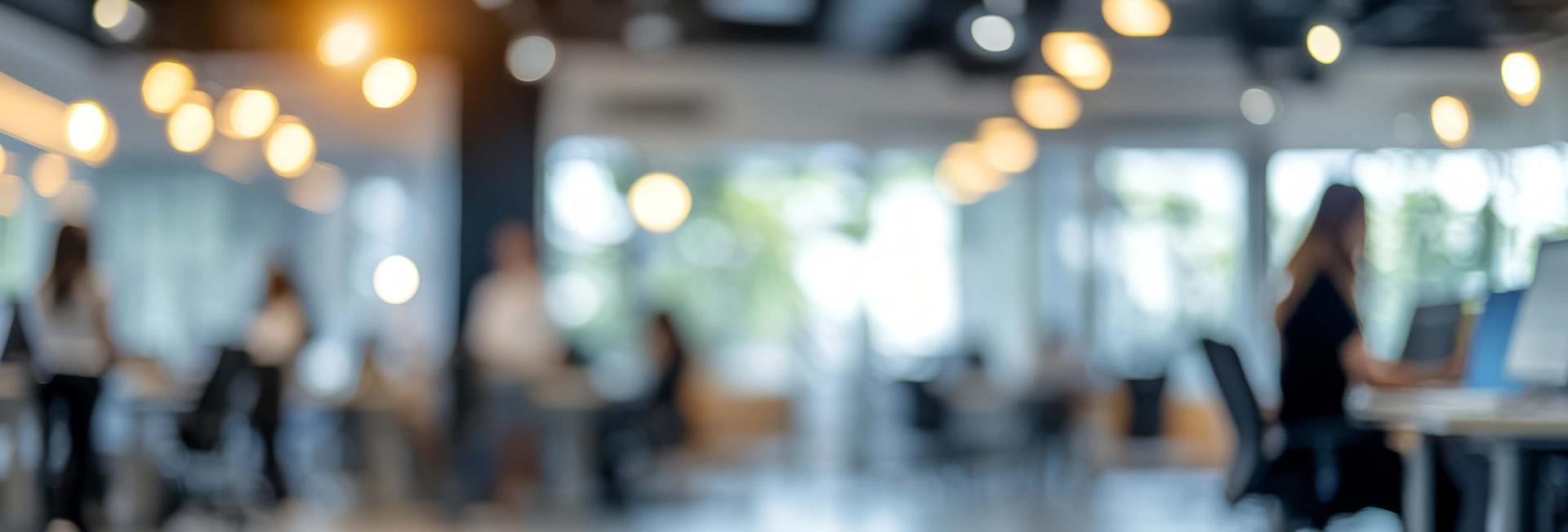 Blurry image depicting individuals seated at desks within an office environment, suggesting a busy workplace atmosphere.