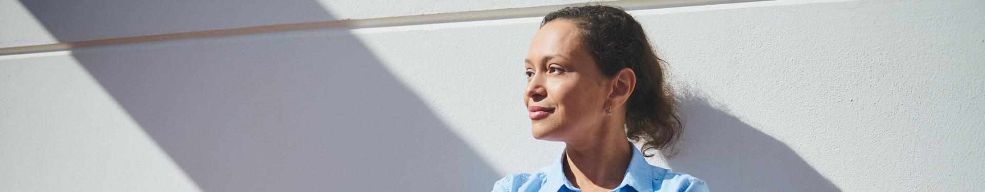 A woman wearing a blue shirt and black pants stands confidently against a clean white wall.