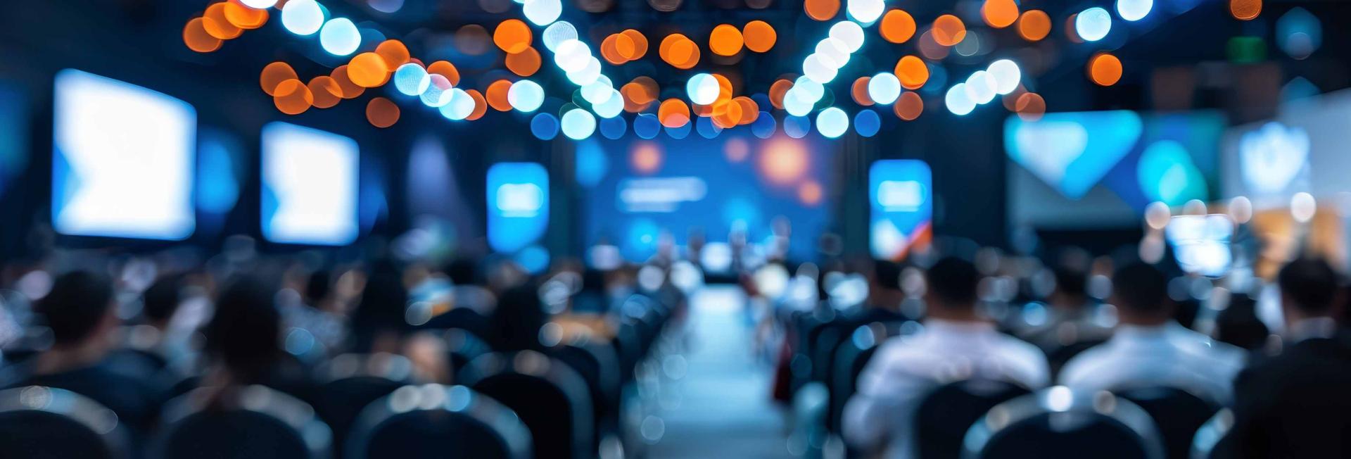 Blurry conference room filled with attendees seated in chairs, engaged in discussion or presentation.