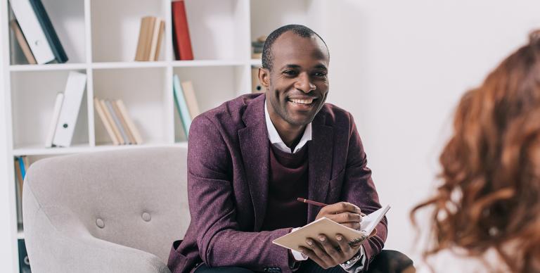 A cheerful man sitting on a chair, smiling as he holds a notepad, conveying a positive and engaging demeanour.