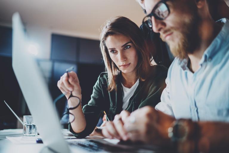 A man and woman are seated together, intently focused on a laptop screen, sharing a moment of collaboration.