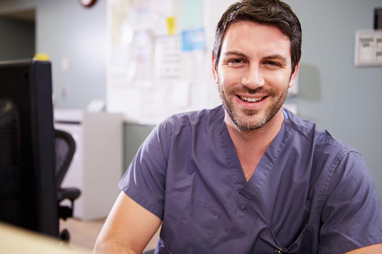A cheerful male mental health student wearing scrubs is seated at a desk, radiating warmth and confidence in his medical environment