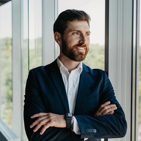 A bearded business law student in a suit stands confidently in front of a large window, showcasing a professional demeanour
