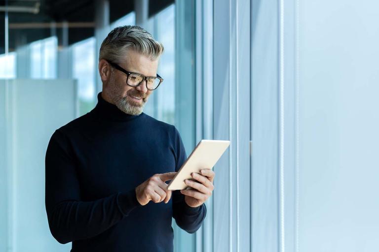 A middle aged MBA student on a tablet smiling in front of a modern office window