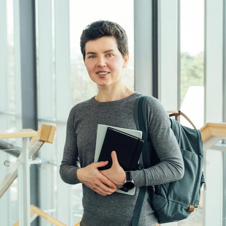 A woman with a backpack and books stands in a hallway, ready to engage in her academic pursuits.
