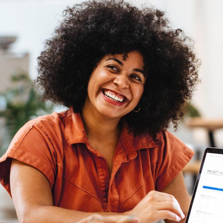 A woman with afro hair smiles brightly while working on her laptop, exuding joy and engagement in her task.