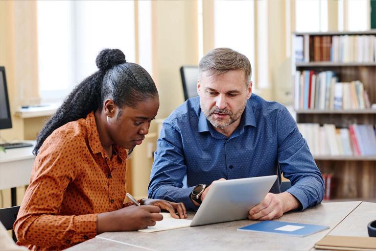 A man and woman seated at a table, engaged with a tablet device between them, sharing ideas or information.