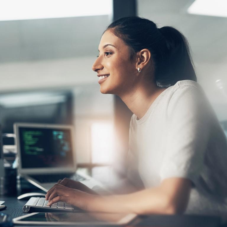 A woman seated at a desk, focused on her laptop, surrounded by a tidy workspace and soft lighting.