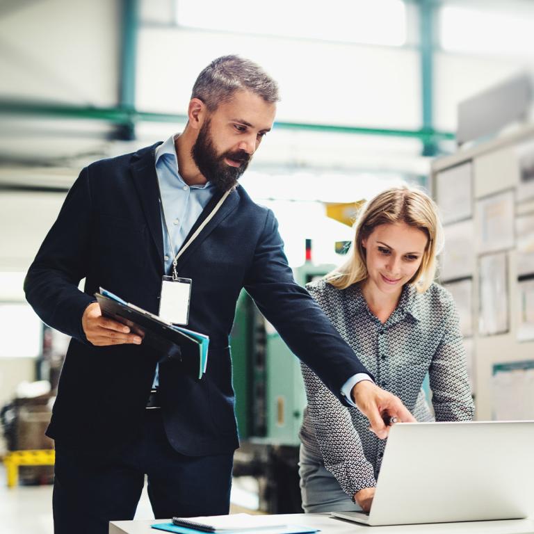A man and woman collaborate on a laptop in a factory setting, focused on their work amidst industrial surroundings.