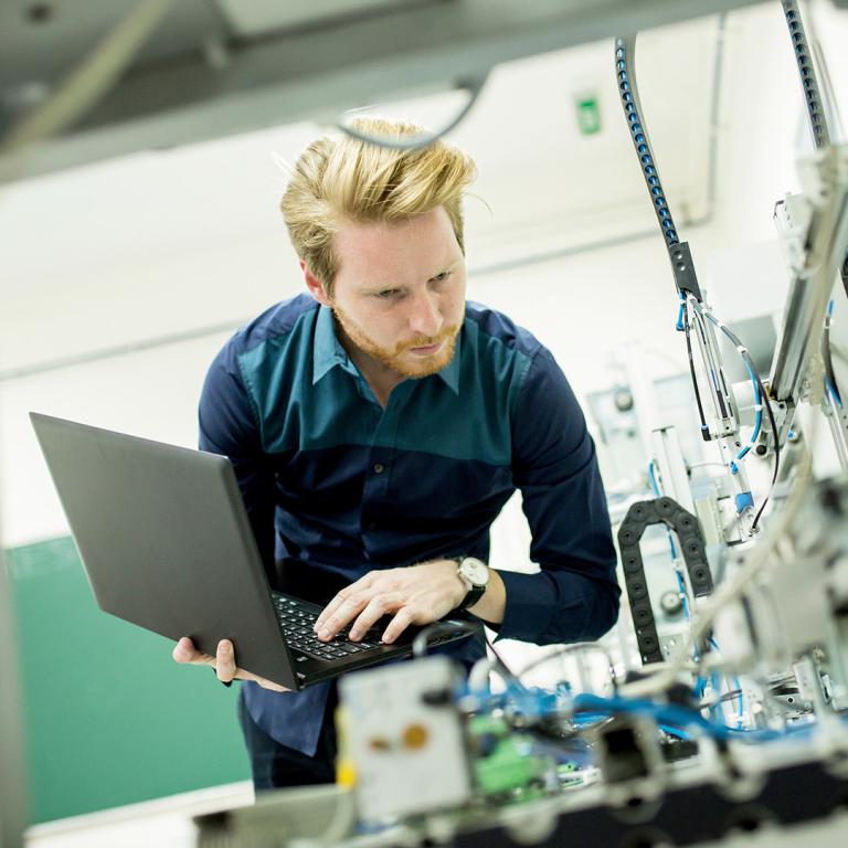 A man focused on his laptop while seated at a workstation in a factory environment.