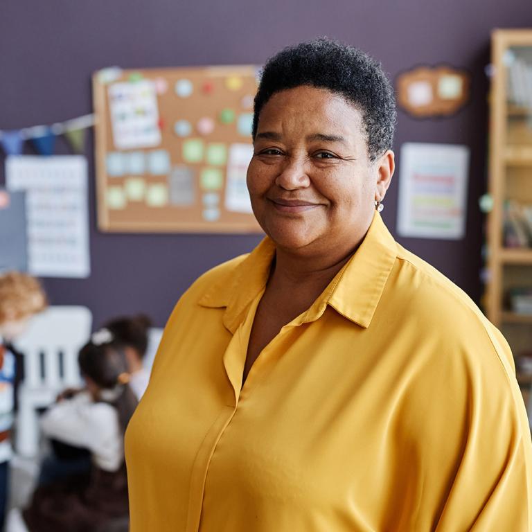 A woman in a yellow shirt stands confidently in front of a classroom, ready to engage with her students.